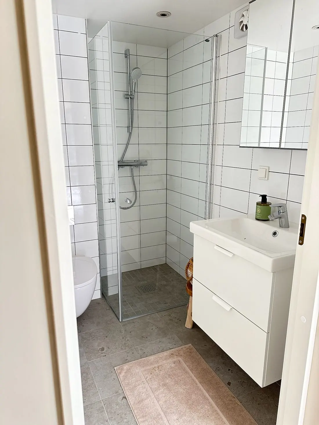 Litet badrum med vitt kakel, dusch bakom glasvägg, vägghängd toalett och tvättställ i ett levererat Öbo-hus på 30 kvm