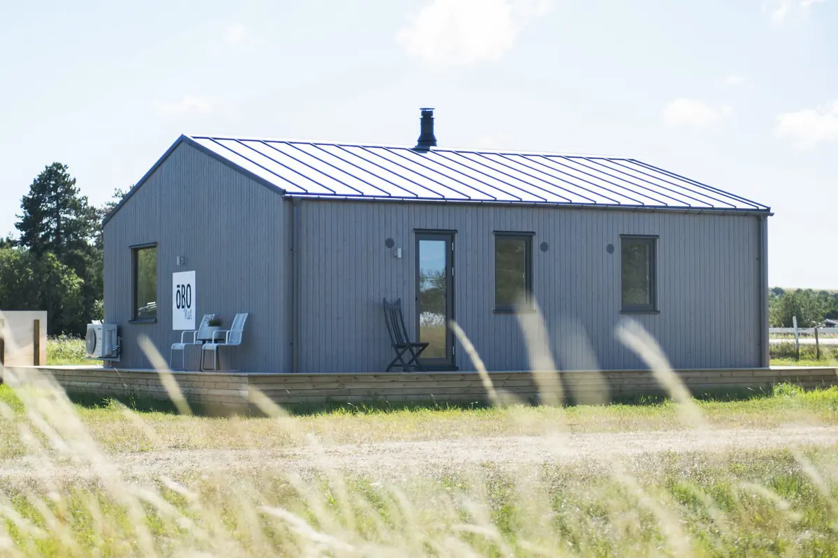 Fårö 74, ett levererat grått trähus med sadeltak och skorsten, sett från baksidan genom högt gräs